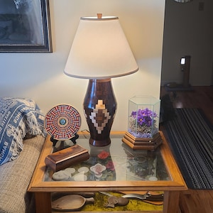 May include: A wooden table lamp with a beige lampshade and a dark brown base with a geometric design. A decorative plate, a wooden box, and a terrarium with purple flowers sit on the table. The table has a glass top.