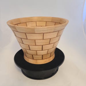 May include: A wooden bowl with a brick pattern design. The bowl is made of light wood and has a dark brown base.