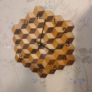 May include: A geometric wooden wall clock with a cube design. The clock features alternating light and dark wood blocks, forming a three-dimensional pattern. Black numerals mark the hours, and black hands indicate the time. The clock is approximately 12 inches in diameter.