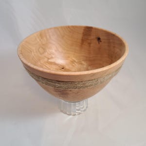 May include: A handcrafted wooden bowl with a light brown finish, accented with a band of natural twine. The bowl sits atop a clear, textured glass base, creating a unique contrast in textures and materials. The bowl's interior shows the natural wood grain.
