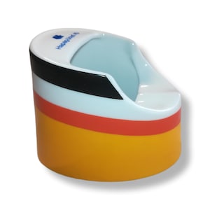 May include: A ceramic ashtray with a curved design. The ashtray features bands of black, white, red, and yellow. The top rim is white with the text "Hapag-Lloyd" in blue. The interior is light blue.