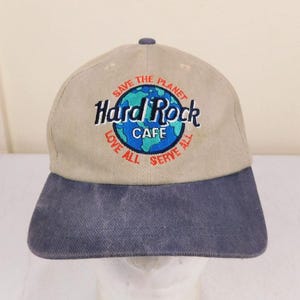 May include: Tan and navy blue Hard Rock Cafe baseball cap. The cap features an embroidered logo with the words "Save the Planet," "Love All," and "Serve All" surrounding a globe design. The Hard Rock Cafe logo is in black and white.