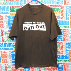 May include: A brown t-shirt featuring the text "When in doubt, Pull Out!" in a white rectangular box. The shirt is hanging against a backdrop with various sports team logos.