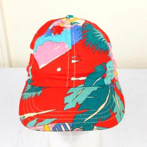 May include: A red baseball cap with a tropical print featuring palm leaves and abstract shapes in teal, pink, blue, and yellow. The cap has a curved brim and a button on top.