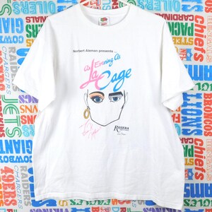 May include: White t-shirt featuring a graphic of a face with blue eyes, gold earring, and the text "An Evening At La Cage" with "Riviera Las Vegas" below. The shirt also has signatures.