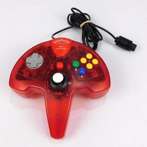 Puede incluir: Un mando SuperPad 64 Colors rojo transparente con un cable negro. El mando tiene una cruceta gris, un joystick blanco y botones de colores etiquetados A, B y C. El texto "SuperPad 64 Colors" es visible.