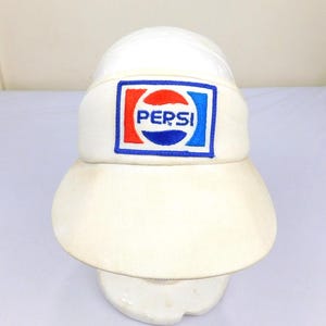 May include: A white visor with the Pepsi logo embroidered on the front. The logo features the word "PEPSI" in white letters inside a red, white, and blue design. The visor has a curved brim and a white band.