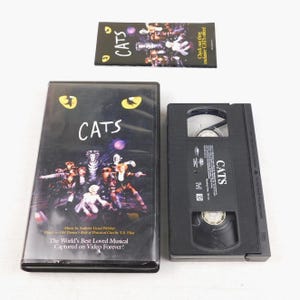 May include: A black VHS tape and case for the musical "Cats" are displayed. The case features the title "Cats" in white, with a cast photo and text. A promotional card with the title "Cats" is also present.
