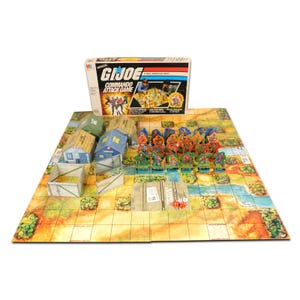 May include: A vintage G.I. Joe Commando Attack Game, featuring a colorful game board with miniature buildings, figurines, and dice. The game box is visible, displaying the game's title and imagery. The game board has a desert landscape.