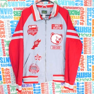 May include: A gray and red sports jacket with multiple patches. The jacket features red sleeves, a gray body, and a white collar. Patches include a bear, basketball, and text like "Cornell" and "Sports Collection".