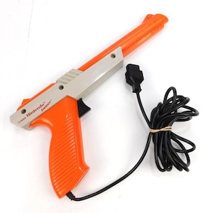 May include: A bright orange and light gray Nintendo Zapper light gun controller with a black trigger and a long black cord ending in a rectangular connector. The side of the device features the text 1985 Nintendo Zapper in red lettering.