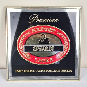 May include: A framed sign for Swan Lager, featuring a black background with gold lettering that reads "Premium" and "Imported Australian Beer." The central design is a red and gold oval with the words "Export Swan Lager" and a swan silhouette.
