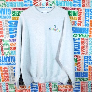 May include: A light gray sweatshirt with the embroidered text "1 Grandma" in colorful letters. The sweatshirt has a crew neck and long sleeves. The background features a colorful pattern of various sports team logos.