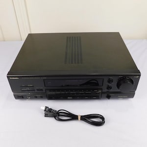 May include: A black Technics SA-G900 stereo receiver with a CD player, various buttons, and a large volume knob. The receiver has a power cord and is on a white surface.
