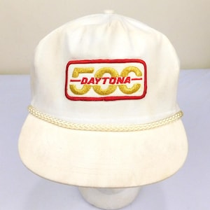 May include: A white baseball cap with a curved brim and a braided rope detail. The cap features an embroidered patch with the text "500 DAYTONA" in gold and red. The cap is a classic style.