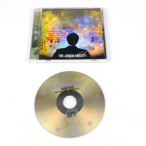 May include: A CD case and disc featuring the album "Wide Eyed" by The Junior Varsity. The case has a colorful, abstract design with a silhouette. The disc is gold with text including "In Stores July 12".