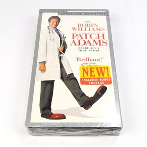 May include: A sealed VHS tape of the film Patch Adams starring Robin Williams. The cover features the actor in a white lab coat and grey trousers, wearing large red clown shoes. Text includes Universal Special Edition, Patch Adams, Based on a True Story, and a yellow sticker stating New! Includes Bonus Footage.