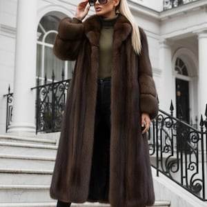 May include: A full-length brown fur coat with a large collar and cuffs. The coat is paired with black trousers and boots. The person is wearing sunglasses and standing on marble steps in front of a white building.