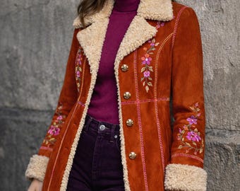 May include: Rust-colored suede coat with floral embroidery and a cream-colored shearling collar and cuffs. The coat features gold buttons and pink stitching details. The model is wearing a burgundy turtleneck and purple jeans.