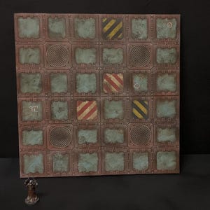 May include: A square game board with a grid pattern, showing a weathered, industrial design. The squares feature various patterns, including diagonal stripes in red and yellow, and circular designs. The board has a rusty aesthetic, with teal and brown colours.