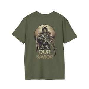 May include: Olive green t-shirt featuring a detailed illustration of a figure in tactical gear holding a rifle. The figure is set against a circular backdrop with the words "OUR SAVIOR" in a camouflage style font.