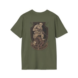 May include: Olive green t-shirt featuring a detailed graphic of a skeletal soldier with a rifle, accompanied by a dog, within a shield design. The words "DEMON HUNTERS" are arched below the image.