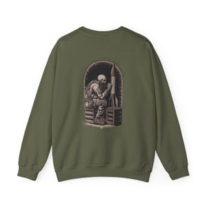 May include: Olive green sweatshirt featuring a detailed graphic on the back. The graphic depicts a skeletal figure in military attire, seated next to a rocket inside an arched frame. The design is rendered in shades of brown and gray.