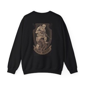May include: Black sweatshirt featuring a detailed graphic on the back. The graphic depicts a soldier with a skull face, holding a rifle, and standing with a dog. The words "DEMON HUNTERS" are printed on a banner below the image.