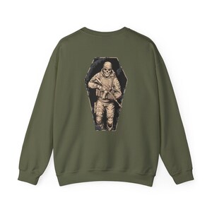 May include: Olive green sweatshirt featuring a detailed graphic on the back. The graphic depicts a skeletal soldier in military gear inside a coffin-shaped frame. The design is in shades of tan and black.
