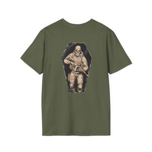 May include: Olive green t-shirt featuring a graphic of a skeletal soldier holding a rifle, set within a coffin-shaped frame. The illustration is detailed with shades of tan and black, creating a distressed, vintage aesthetic. The shirt is a standard crew neck.