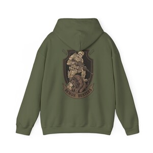 May include: Olive green hoodie featuring a graphic of a soldier with a skull head, holding a rifle, and a snarling creature. The graphic is within a shield shape with the words "Demon Hunters" on a banner.