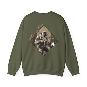 May include: Olive green sweatshirt featuring a diamond-shaped graphic on the back with a soldier illustration. The illustration is in shades of brown and beige, with the words "SPRAY & PRAY" above the graphic.