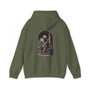 May include: Olive green hoodie featuring a detailed illustration on the back. The graphic depicts a skeleton in military gear seated next to a rocket inside an arched frame. The hoodie has a hood and long sleeves.