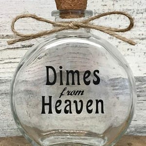 May include: Clear glass jar with a cork stopper and a twine bow. The jar is round with a flat bottom and features the words "Dimes from Heaven" in black lettering. A decorative item.