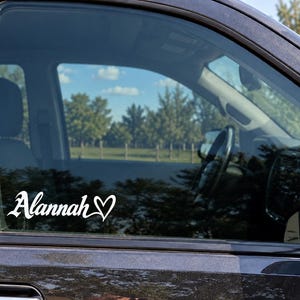 May include: A white vinyl decal applied to the exterior of a dark vehicle window. The decal features the name Alannah written in a stylized, cursive script font, followed by a small, simple heart outline symbol positioned to the right of the text.