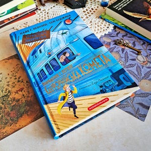 Könnte beinhalten: Ein blaues Hardcover-Buch mit einer Zugillustration auf dem Cover. Der Titel ist in einer Fremdsprache, mit einem Jungen in einem gelben Mantel, der auf dem Cover rennt. Weitere Bücher und dekorative Gegenstände sind im Hintergrund.