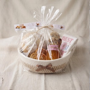 May include: A gift basket filled with baked goods, jam, and a decorative towel. The basket contains a loaf of bread, cookies, dried orange slices, and a jar of jam with a pink and white checkered lid. The basket is wrapped in clear cellophane and tied with twine.