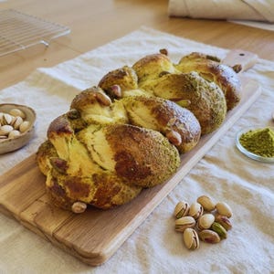 May include: Braided loaf of bread with a golden-brown crust, sprinkled with green pistachio powder and whole pistachios. The bread sits on a wooden cutting board, with a small bowl of pistachios and a dish of green powder nearby.