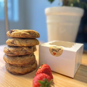 May include: A stack of five chocolate chip cookies next to a white bakery box with a clear window and a sticker that says "Thank you!" with a heart. Two fresh strawberries are in front of the box.