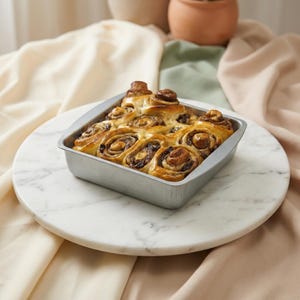 May include: A square, metal baking pan filled with freshly baked cinnamon rolls. The rolls are golden brown with a glossy glaze, and sit on a white marble serving board. The background includes neutral-toned fabrics and pottery.