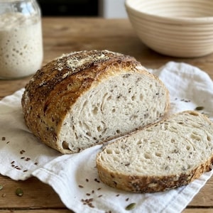 May include: A loaf of freshly baked bread, sliced to reveal the airy interior with visible seeds. The bread rests on a white linen cloth, with a jar of starter and a woven bowl in the background. The bread is a golden brown color.