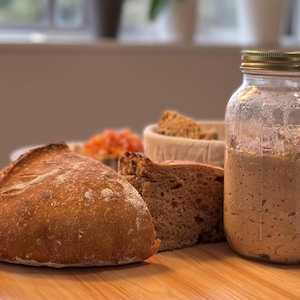 May include: A loaf of golden-brown bread, a slice of bread, and a jar of sourdough starter sit on a wooden surface. A basket with bread pieces and a small dish of orange food are also present. The jar has a gold lid.