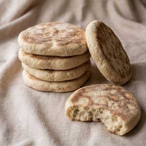 May include: A stack of six round, golden-brown flatbreads, with a slightly textured surface, sits on a beige linen cloth. One flatbread is leaning against the stack, and another has a bite taken out of it, revealing the soft interior.
