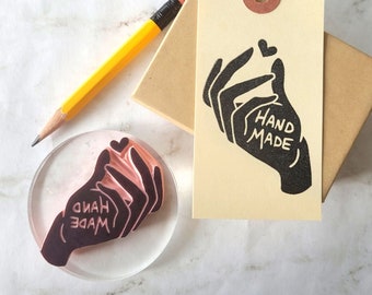 May include: A clear, round stamp with a hand-drawn design that says "HAND MADE" and a matching tag. A yellow pencil and a small brown box are also in the image. The tag has a heart above the hand and is tied with red and white twine.