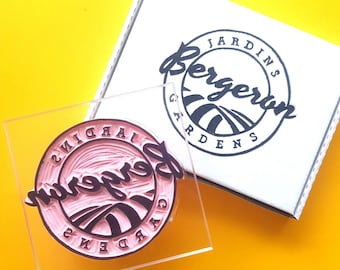May include: A clear acrylic stamp with a pink ink impression of the "Bergeron Gardens" logo, featuring a circular design with text and a stylized landscape. A white box with the same logo is in the background, set against a yellow backdrop.