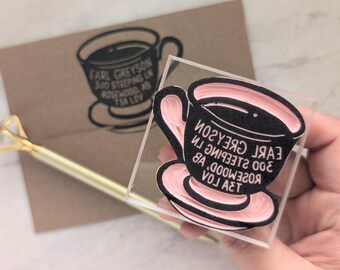 Pode incluir: Um carimbo de borracha acrílico transparente com um design de xícara de chá e o texto EARL GREYSON 300 STEEPING LN ROSEWOOD AB T3A L0V. O carimbo tem uma base rosa e detalhes em borracha preta, exibindo o mesmo design num envelope castanho.