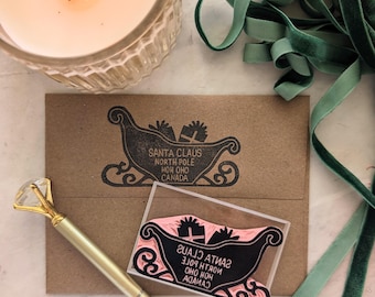 Pode incluir: Um envelope de papel castanho carimbado com tinta preta com um trenó carregado de presentes e o texto SANTA CLAUS NORTH POLE HOH OHO CANADA. Ao lado, um carimbo de borracha rosa com o mesmo design e uma caneta dourada com topo de cristal.