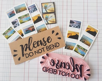 May include: A brown paper card with the text "please do not bend" in black script. The card is surrounded by a collection of Canadian postage stamps with images of landscapes and the word "Canada" printed on them. There is also a pink rubber stamp with the text "please do not bend" in black script.