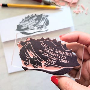 May include: A black and pink rubber stamp with a dragon design and text. The stamp is held up, revealing the mirrored text and image. A stamped image is visible on a white surface in the background.