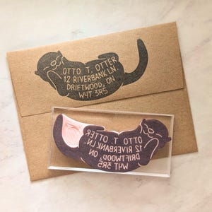 May include: A custom address stamp with an otter design in black ink. The stamp reads "OTTO T. OTTER, 12 RIVERBANK LN., DRIFTWOOD, ON, W4T 3R5". A brown envelope is visible in the background.